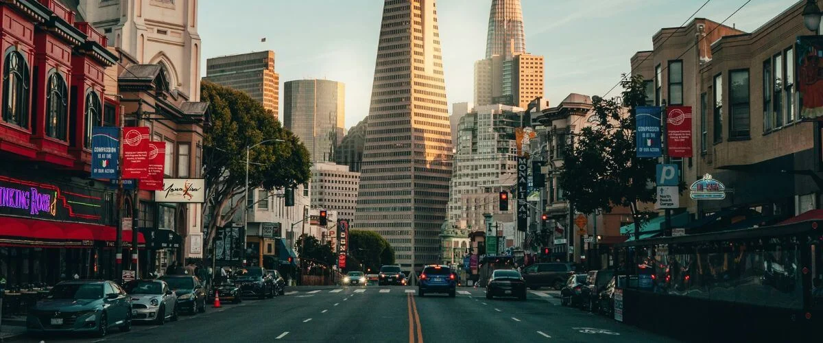 Sunset Street View of San Francisco’s Transamerica Pyramid Cars driving down a busy San Francisco street at sunset, with the Transamerica Pyramid and other downtown skyscrapers rising in the background and colorful storefronts lining both sides of the road.