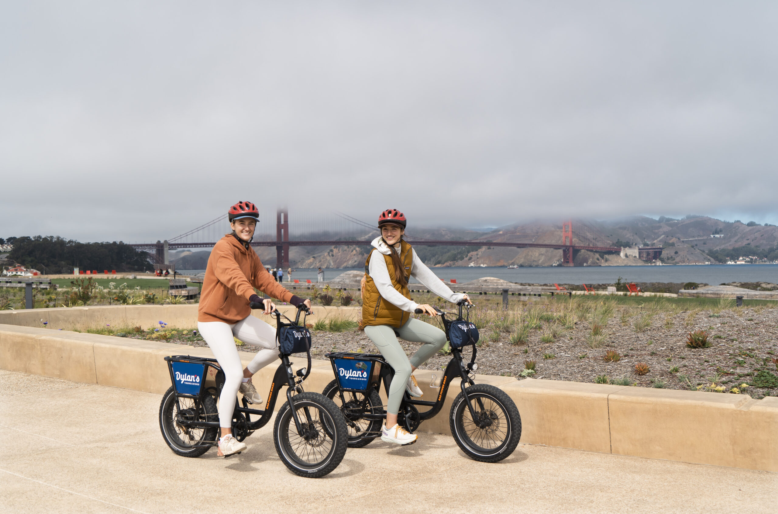 Bikes22 Two people wearing helmets ride e-bikes near the iconic Golden Gate Bridge. Dressed casually and smiling, they enjoy the scenic waterfront view on a cloudy day, embracing the adventure in style.