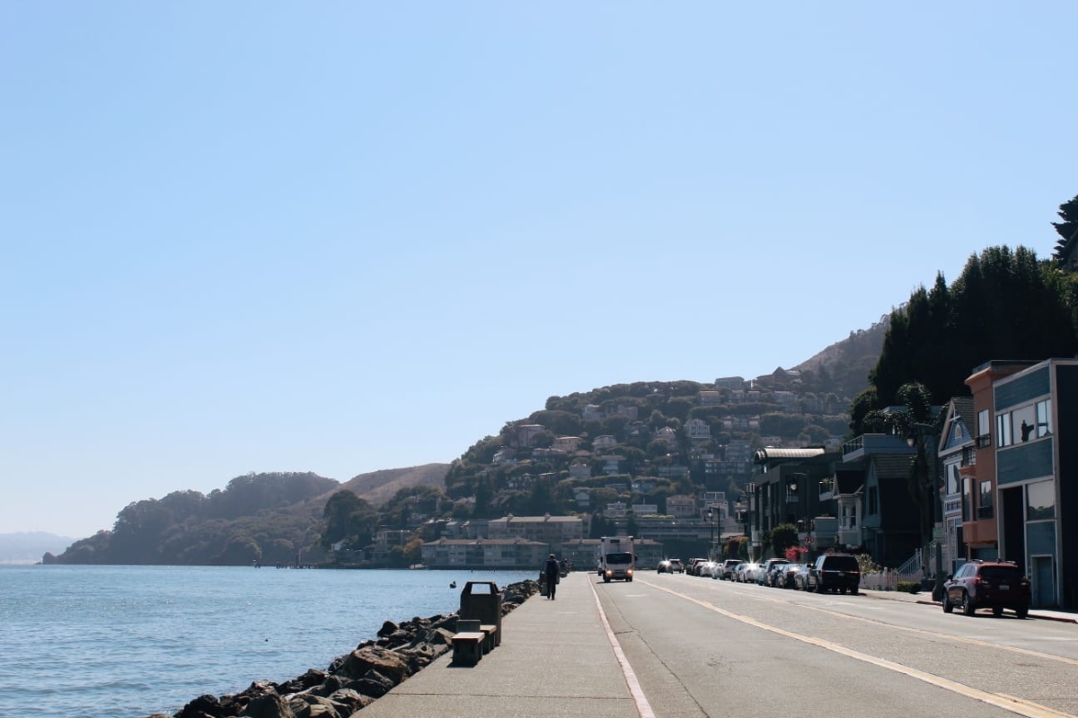 Waterfront road in Sausalito with rocky shoreline, parked cars, and hillside homes overlooking San Francisco Bay