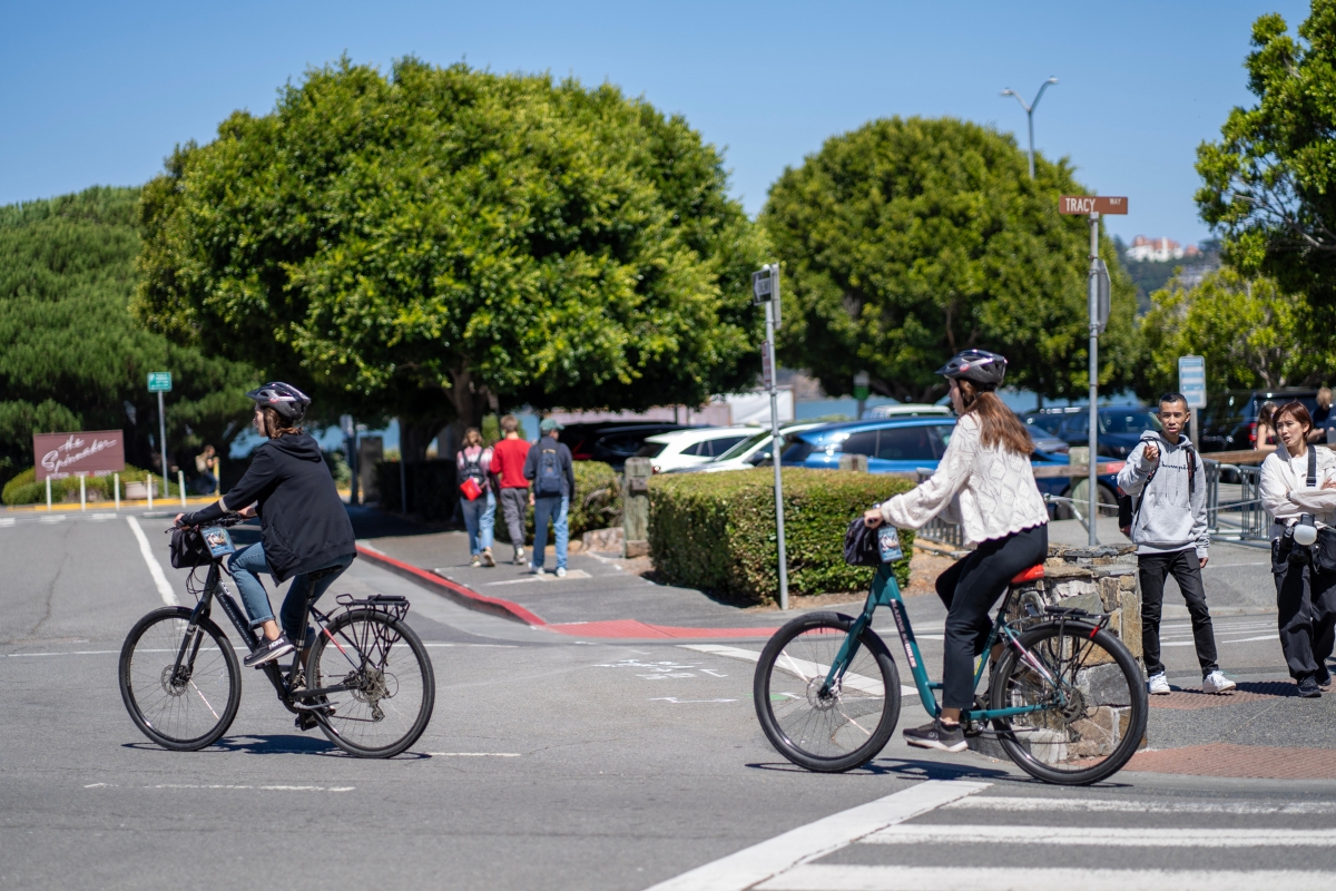 Two cyclists riding near Tracy Way intersection with lush trees, bike lanes, and pedestrians in Marin County
