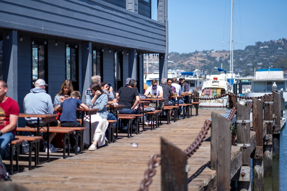 People enjoying outdoor dining on wooden deck at waterfront restaurant with boats and hills in background