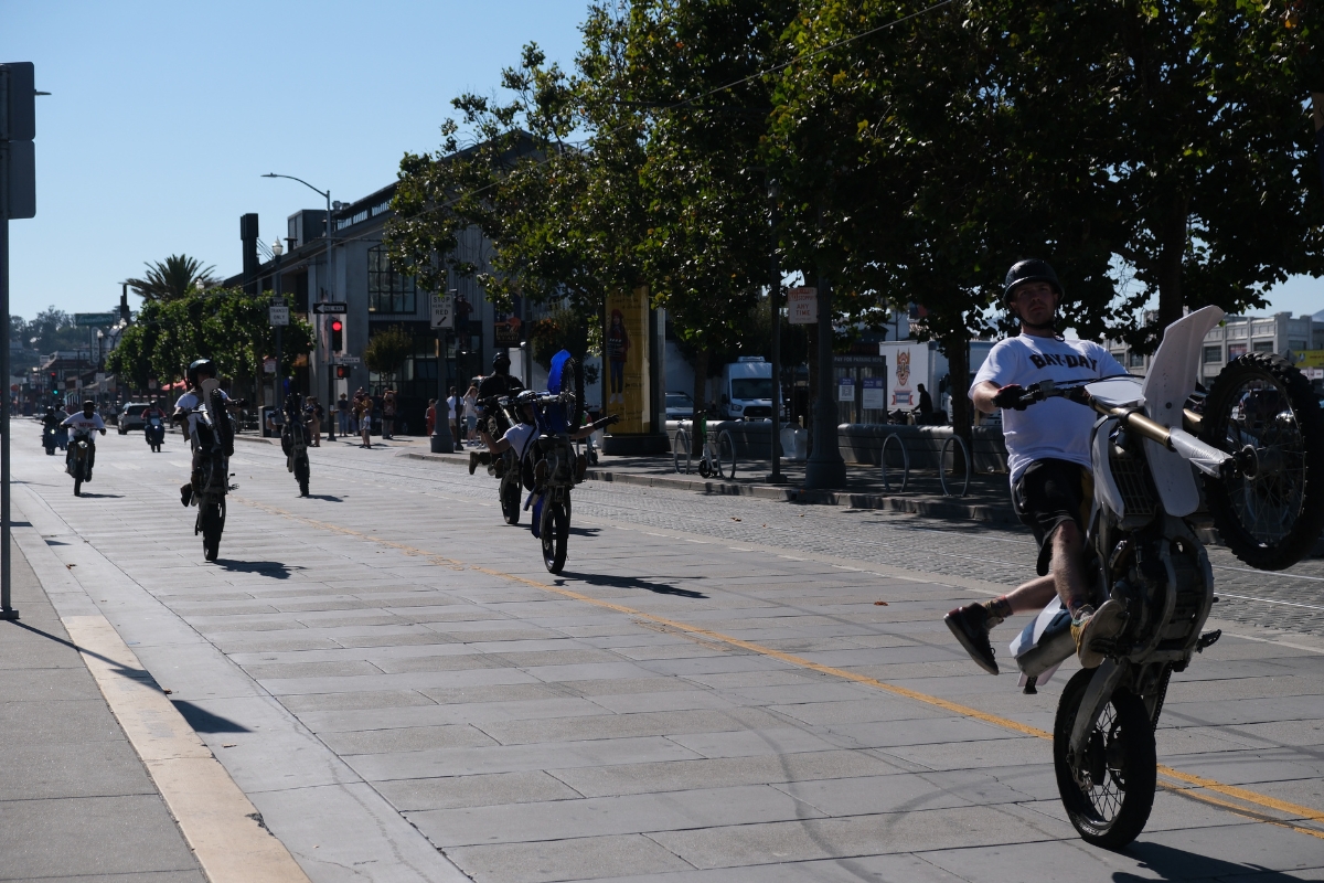 Motorcycle riders performing wheelies on a paved street near Fisherman's Wharf with shops and trees in background.