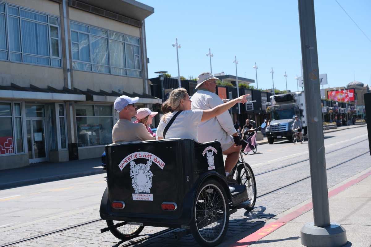 Family enjoying a pedicab tour on a sunny day in San Francisco, riding in a Sweatie's Art Bar rickshaw along a street with trolley tracks and shops
