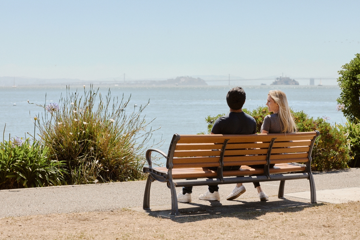 Couple sitting on wooden bench overlooking bay with distant Golden Gate Bridge, Alcatraz Island, and coastal vegetation