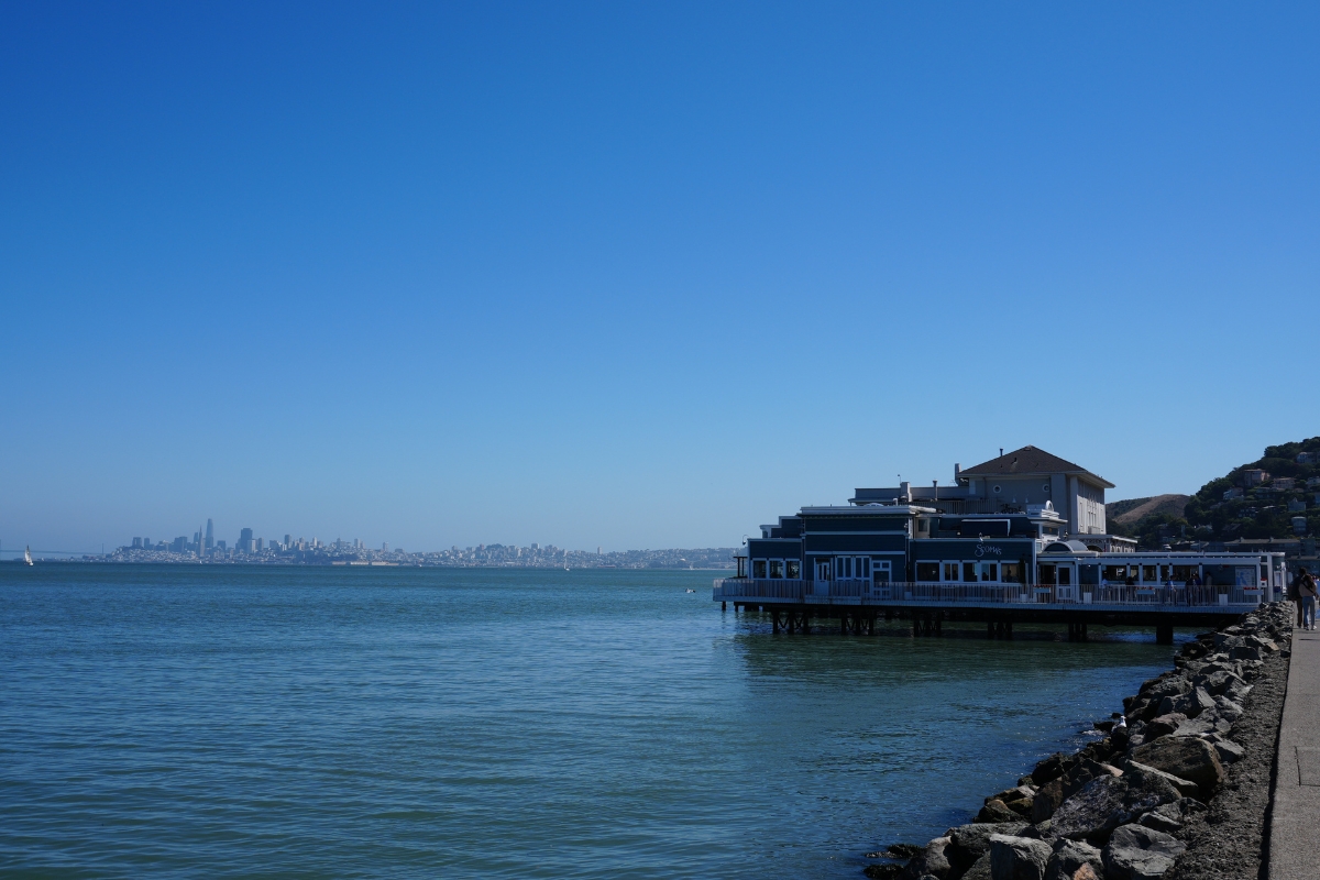 Blue restaurant building on stilts over water with city skyline visible across the bay