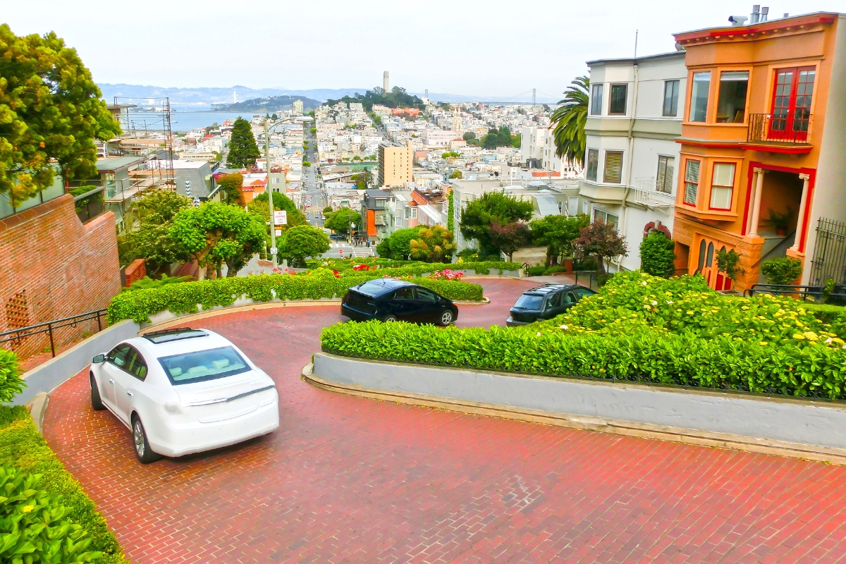 Aerial view of Lombard Street's iconic winding road with flowerbeds, parked cars, and panoramic views of San Francisco cityscape, Coit Tower, and the Bay Bridge.