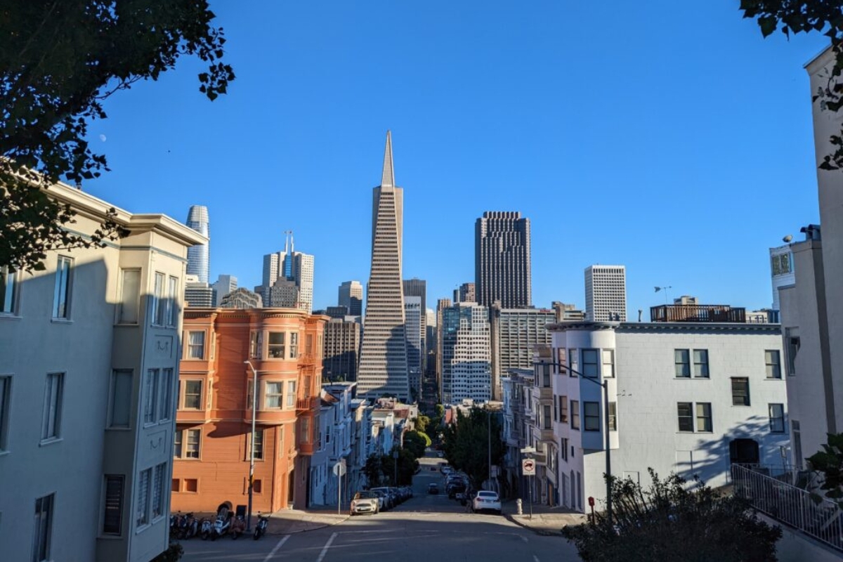 View of San Francisco skyline featuring Transamerica Pyramid and downtown skyscrapers seen from steep residential street with colorful Victorian buildings