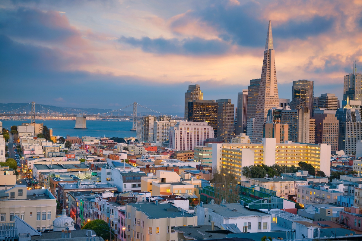 Panoramic view of San Francisco skyline featuring Transamerica Pyramid, downtown skyscrapers, Bay Bridge, and colorful residential buildings at sunset