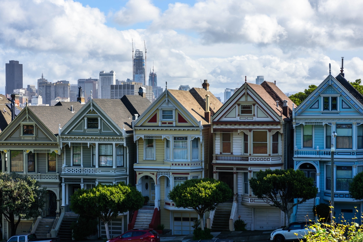 Row of colorful Victorian Painted Ladies houses with downtown San Francisco skyline in background under blue sky with clouds
