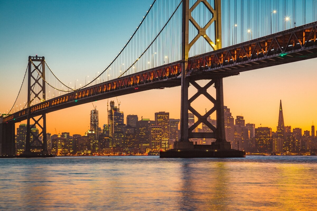 San Francisco-Oakland Bay Bridge lit up at sunset with downtown San Francisco skyline silhouetted against orange sky