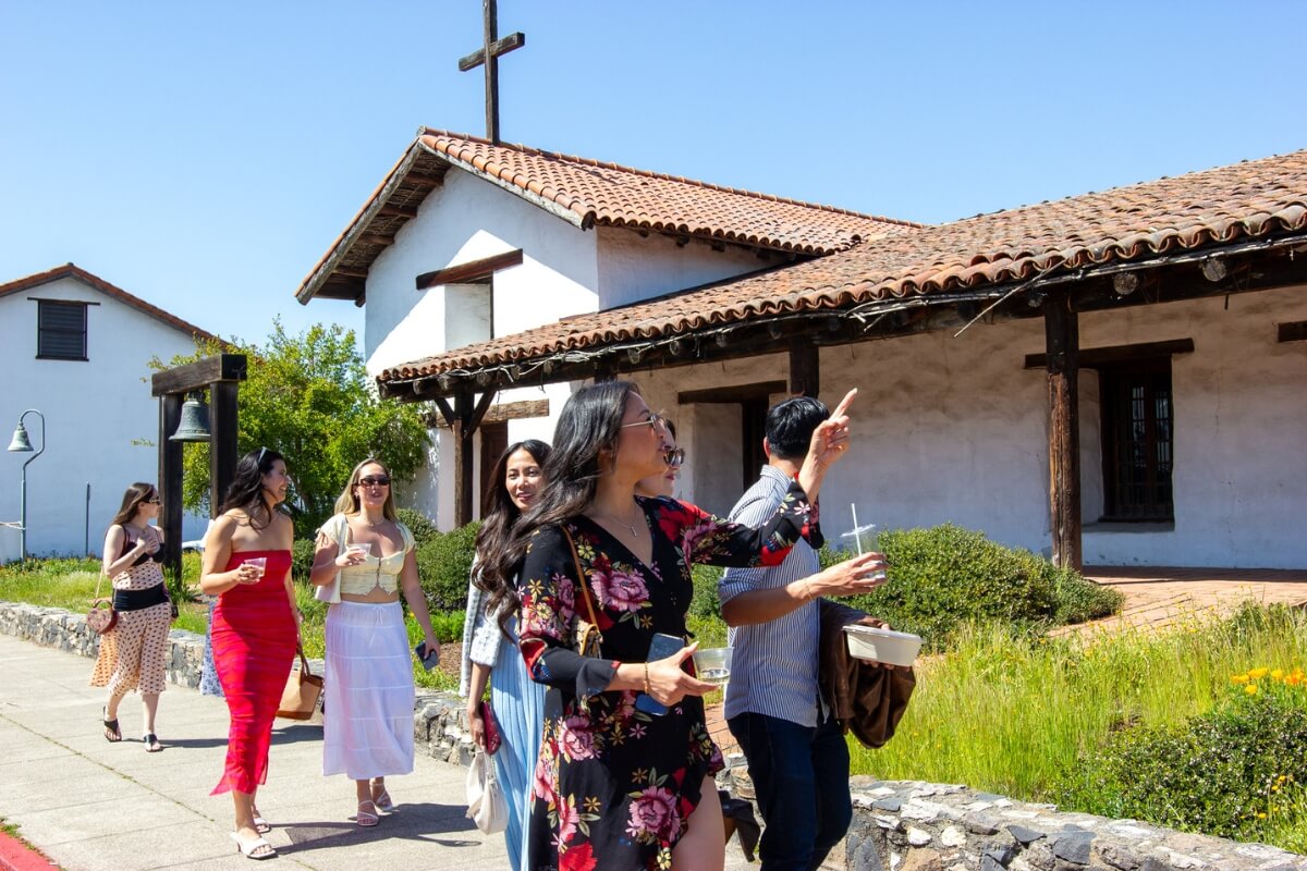 Group touring Spanish colonial mission with terra cotta roof and cross in California wine country on sunny spring day.