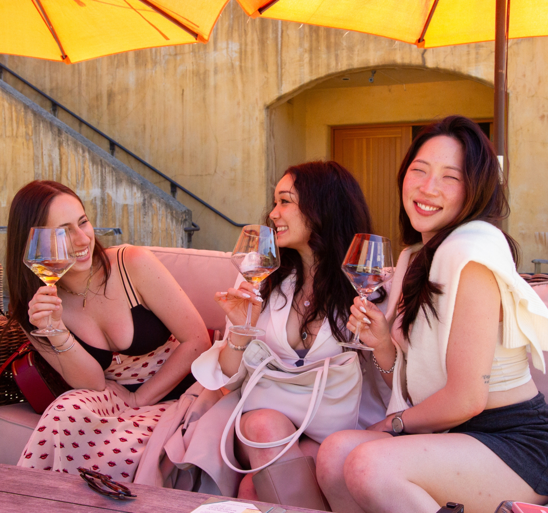 Three women enjoying wine glasses on an outdoor patio under orange umbrellas during Dylan's Famous Wine Country Tour in Napa Valley