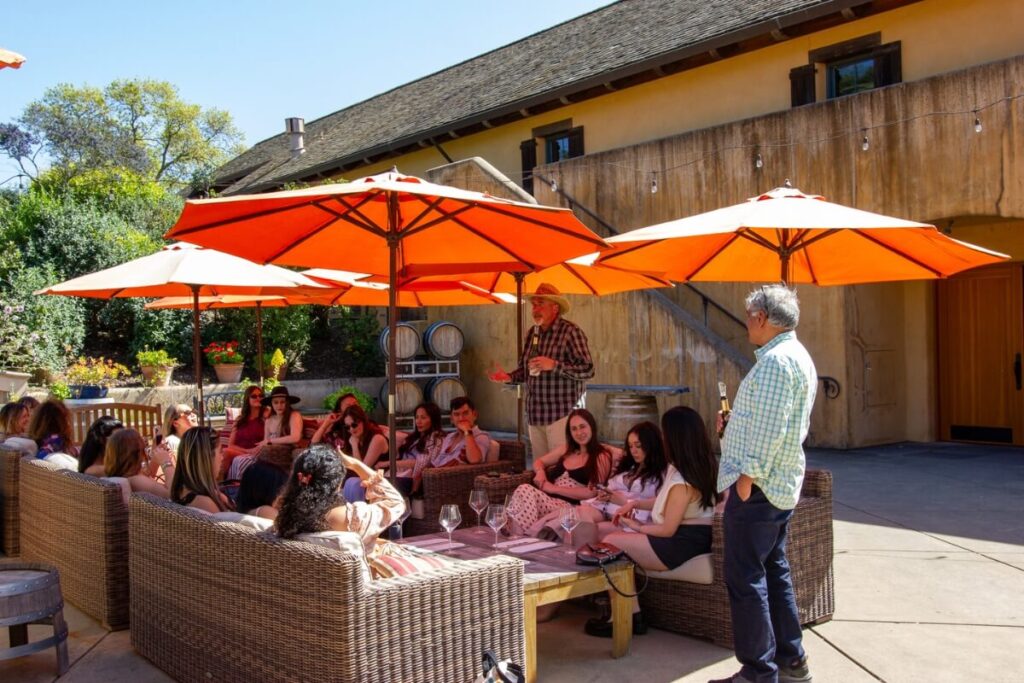 Wine country tour group relaxing on outdoor patio with orange umbrellas while winery staff conducts guided tasting session near barrel storage