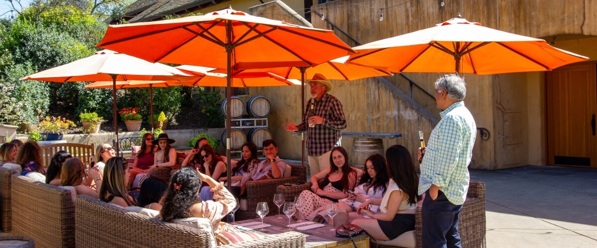 Winery guests relaxing under bright orange umbrellas during outdoor wine tasting session with vineyard staff in California wine country