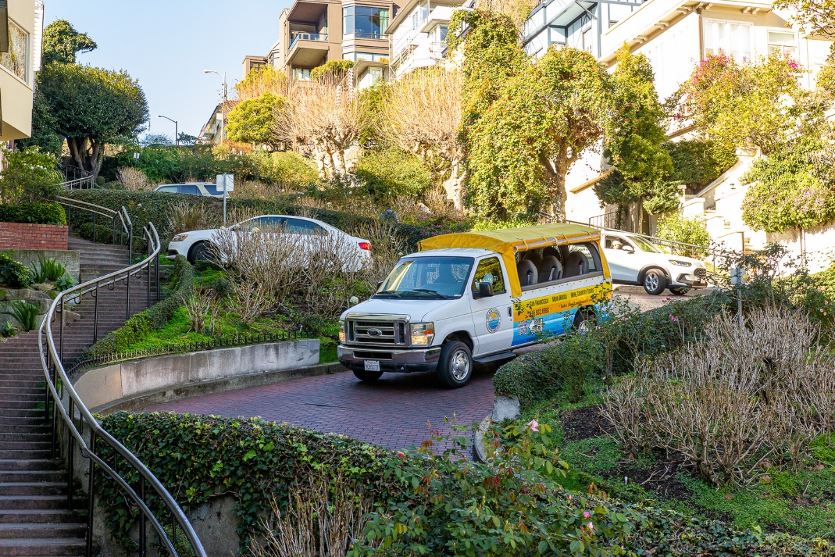 White and yellow Dylan's Tours van with blue trim driving on a brick pathway surrounded by lush landscaping and hillside apartment buildings in San Francisco