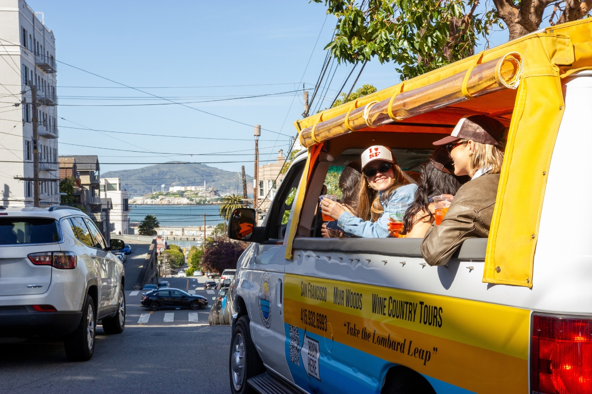 Dylan's Tours van with tourists looking out at San Francisco Bay view, showing Golden Gate Bridge in distance and city streets below