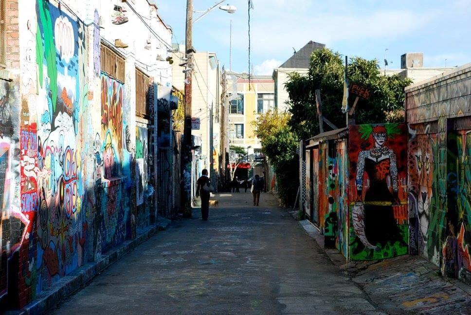 Narrow alley in San Francisco’s Mission District lined with vibrant murals on both sides, with a few people walking toward the sunlit end.