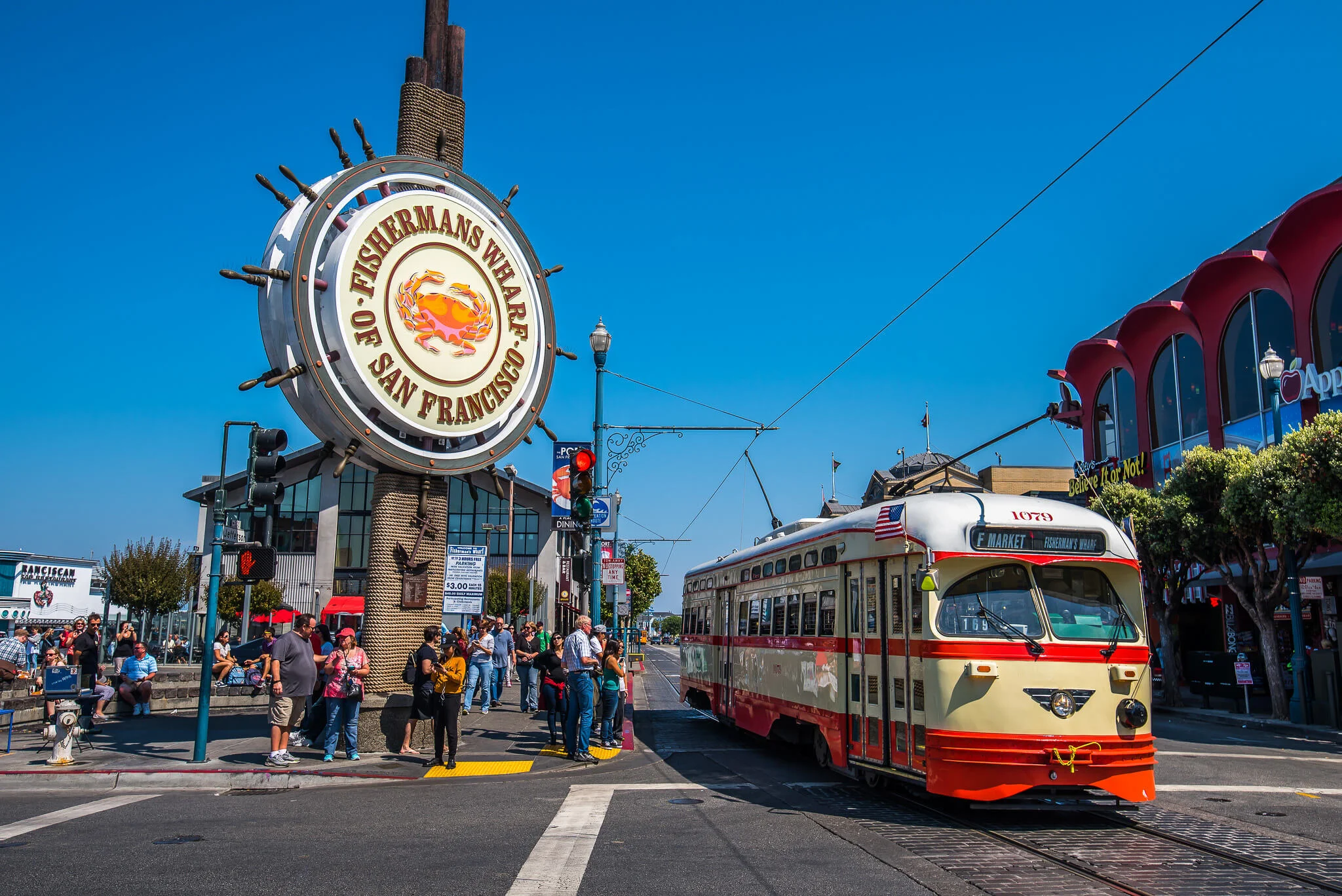 The large Fisherman’s Wharf of San Francisco sign towers over a busy corner as a red-and-cream historic F-Market streetcar pulls in, with crowds waiting under a clear blue sky.