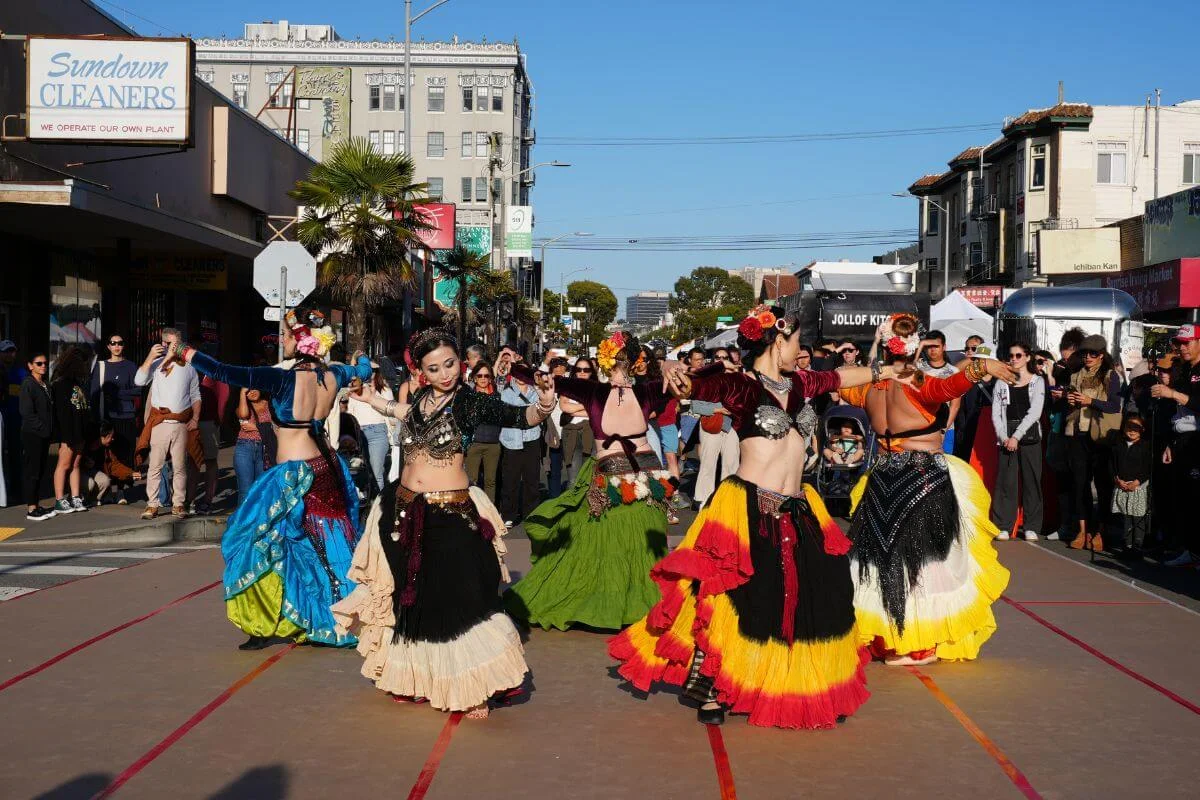 Group of dancers in bright, layered skirts performing a belly-dance–style routine in the middle of a closed-off street, surrounded by a crowd of onlookers and food trucks on a sunny day.
