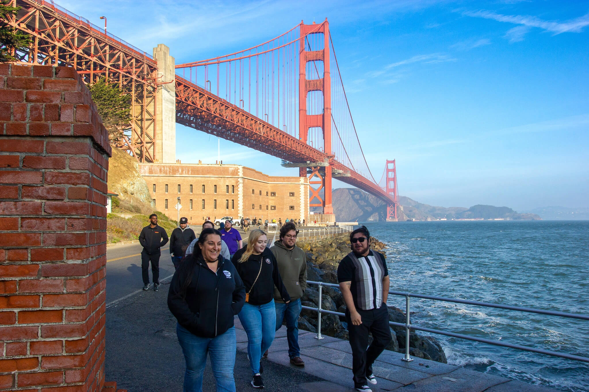 Group of people walking along the waterfront by Fort Point, with the Golden Gate Bridge stretching overhead and the bay on a clear blue day.