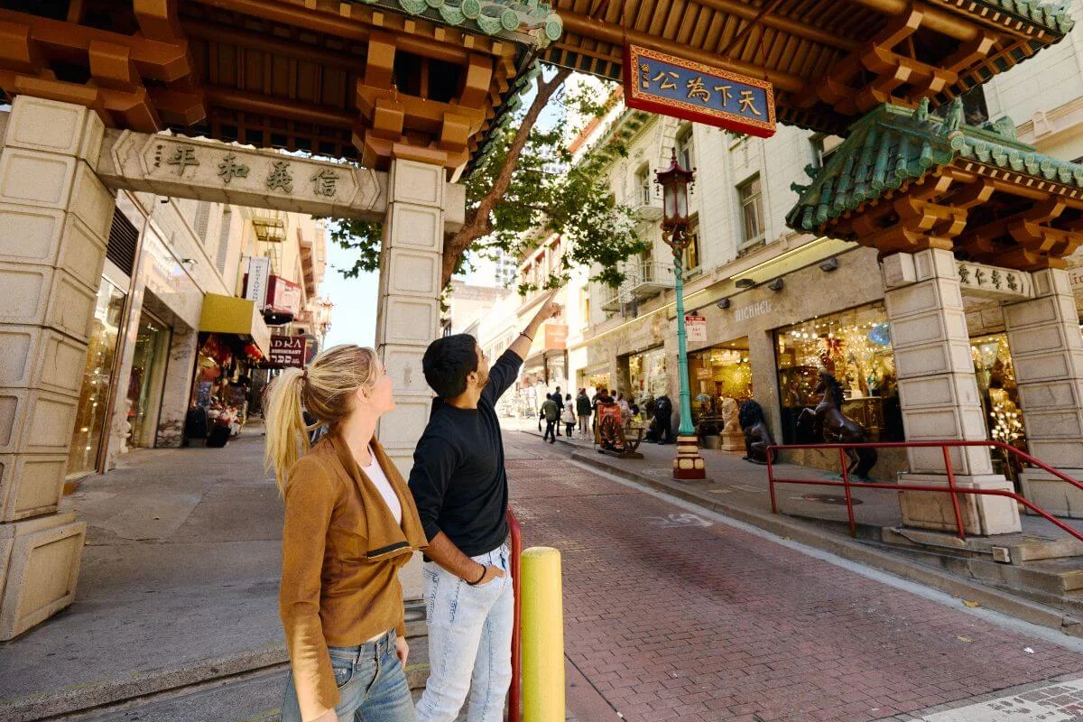 Two visitors beneath the Dragon Gate on Grant Avenue, looking up at the ornate arch as they enter Chinatown’s shop-lined street on a sunny day.