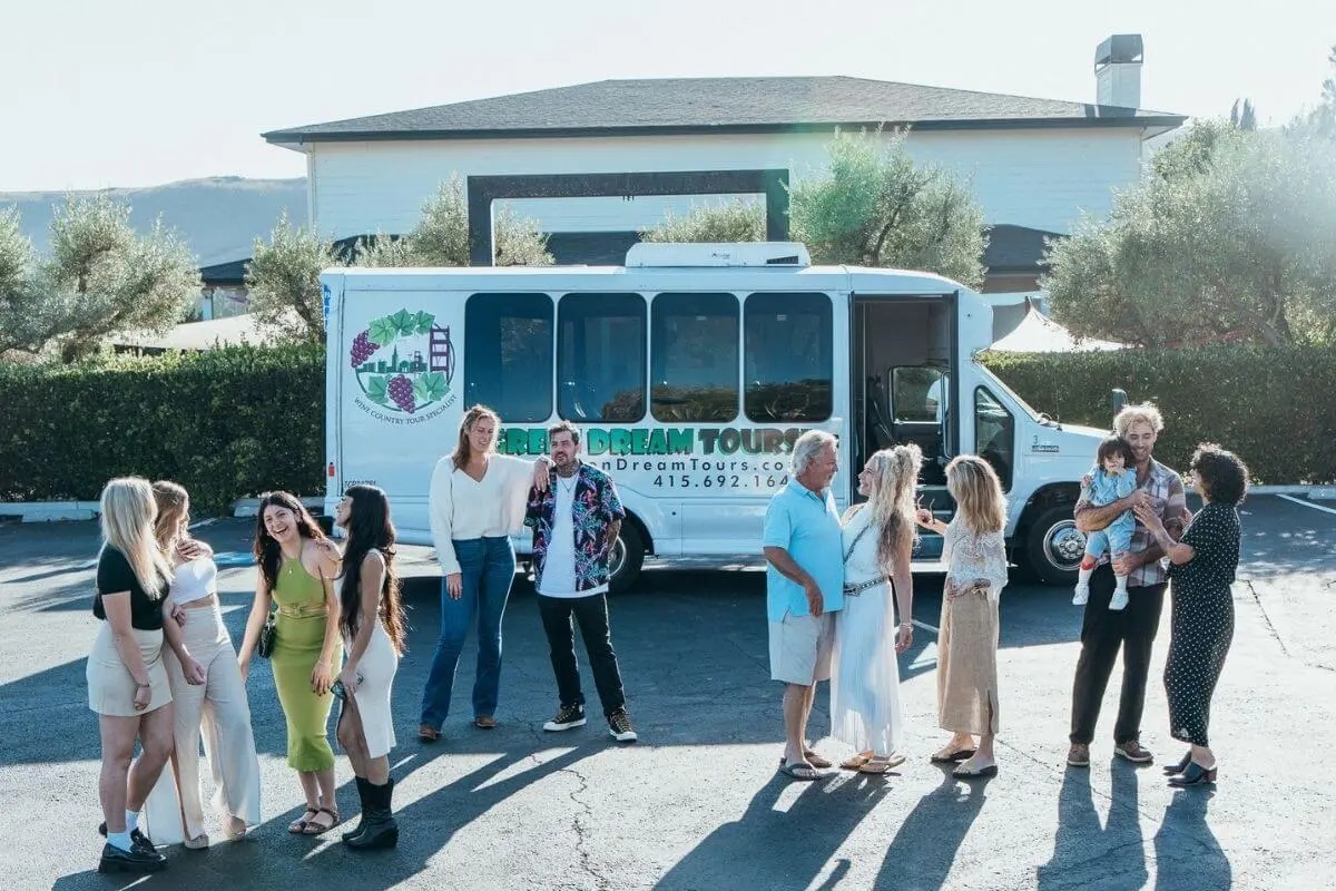 A group of people stand and chat in a sunny parking lot beside a white tour shuttle labeled “Green Dream Tours,” preparing for a wine country tour. The group includes men, women, and a child, all dressed casually and smiling in front of a backdrop of greenery and a modern building.