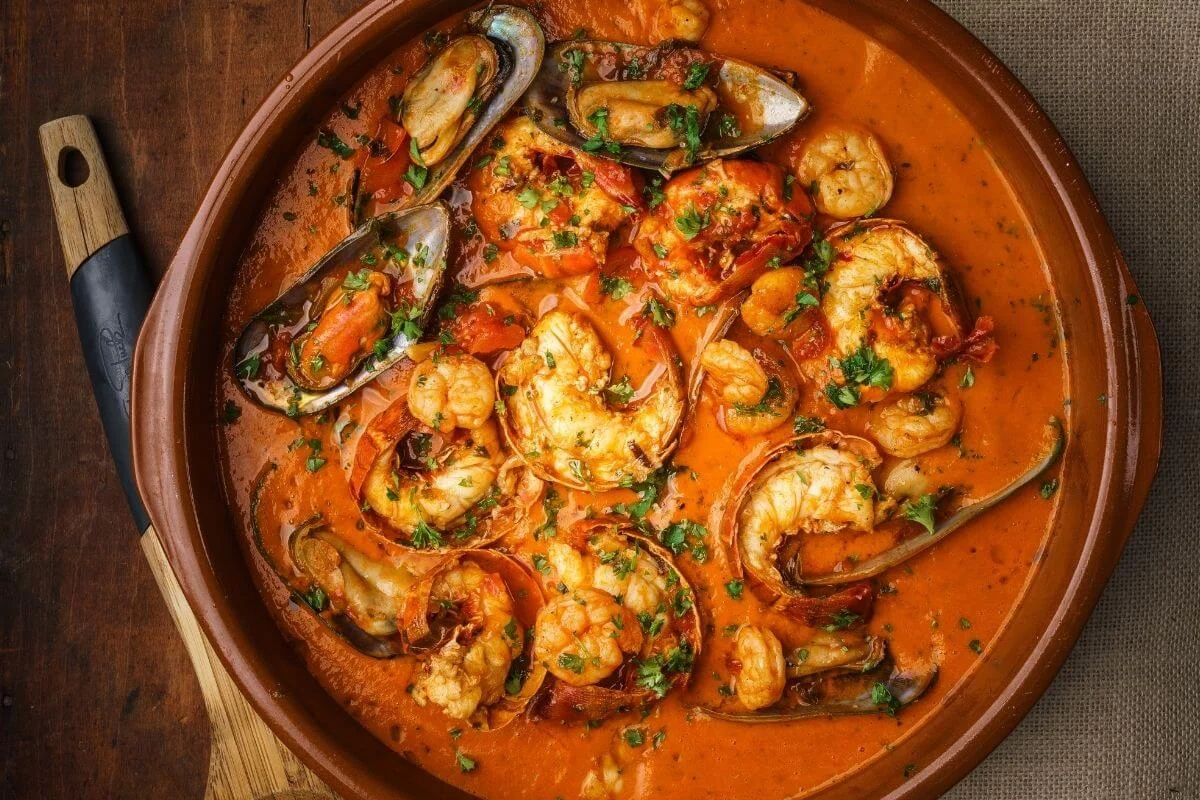 Overhead view of a rustic clay pot filled with rich tomato-based seafood stew, featuring shrimp, mussels, and other shellfish garnished with chopped fresh herbs on a wooden board.