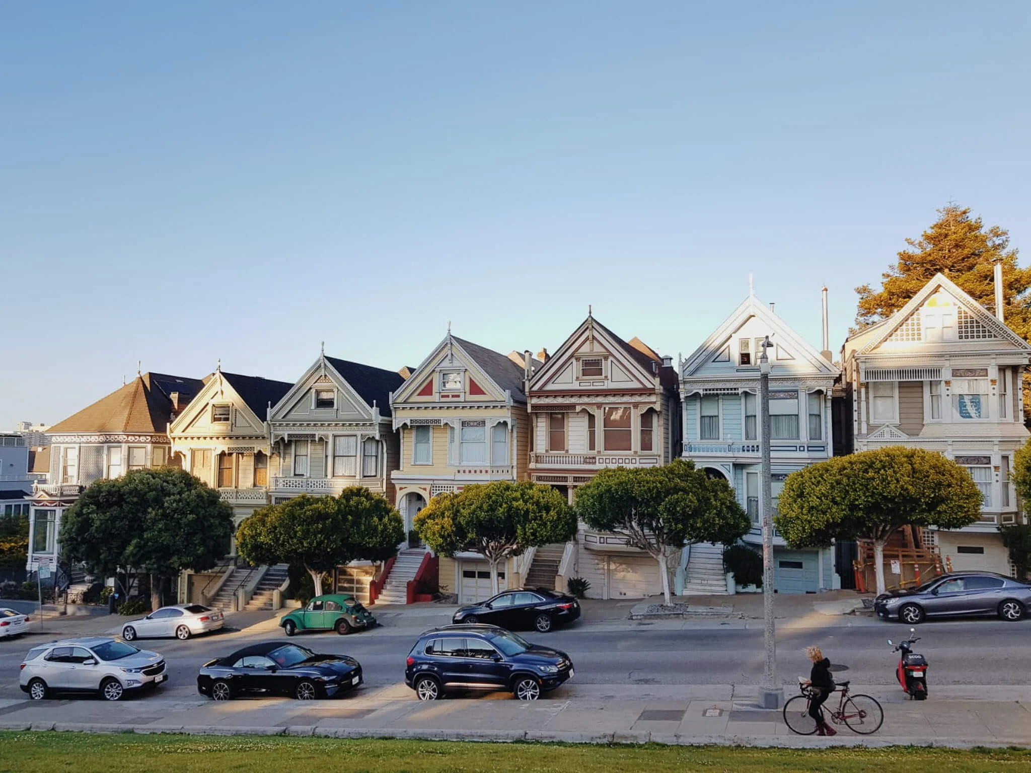 Wide view of the iconic Painted Ladies Victorian houses lined up along a street with parked cars, trimmed trees, and a cyclist passing by under a clear evening sky.