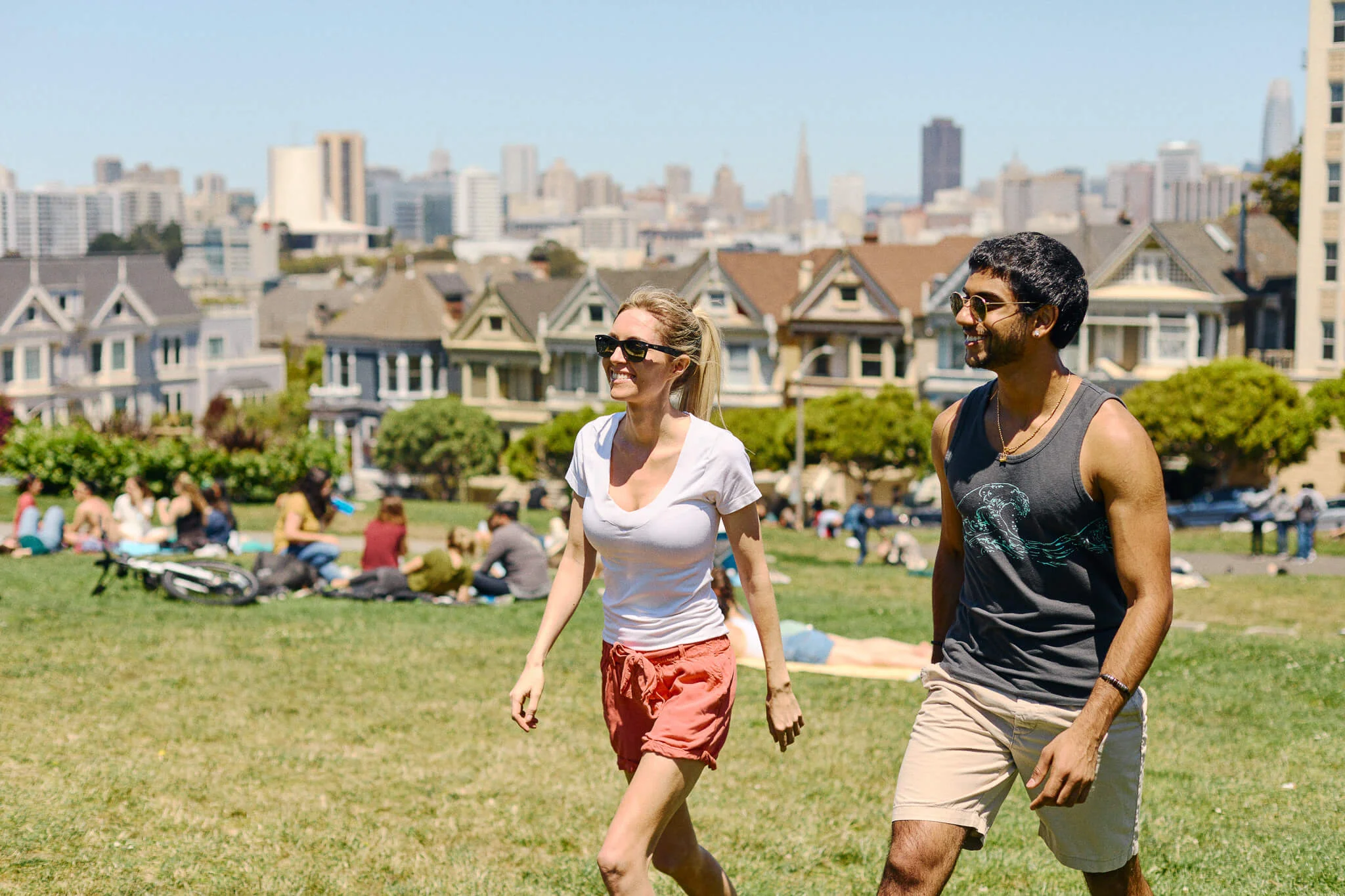 Smiling man and woman walk across a sunny grassy park at Alamo Square, with people relaxing on the lawn and the Painted Ladies houses and San Francisco skyline in the background.