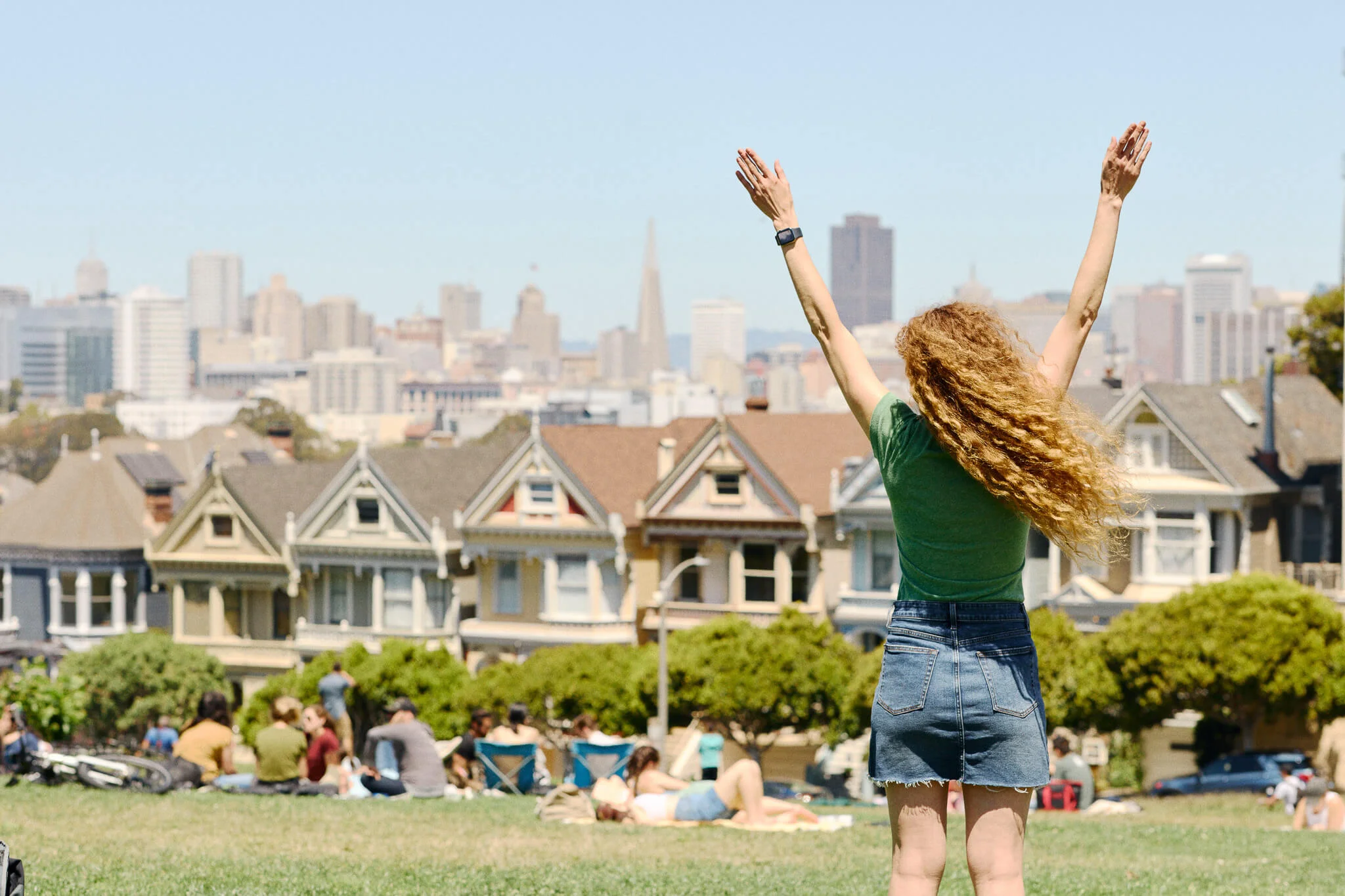 Rear view of a woman with long curly hair raising her arms in the air while standing in Alamo Square Park, with people relaxing on the grass, the Painted Ladies houses, and the San Francisco skyline in the background.
