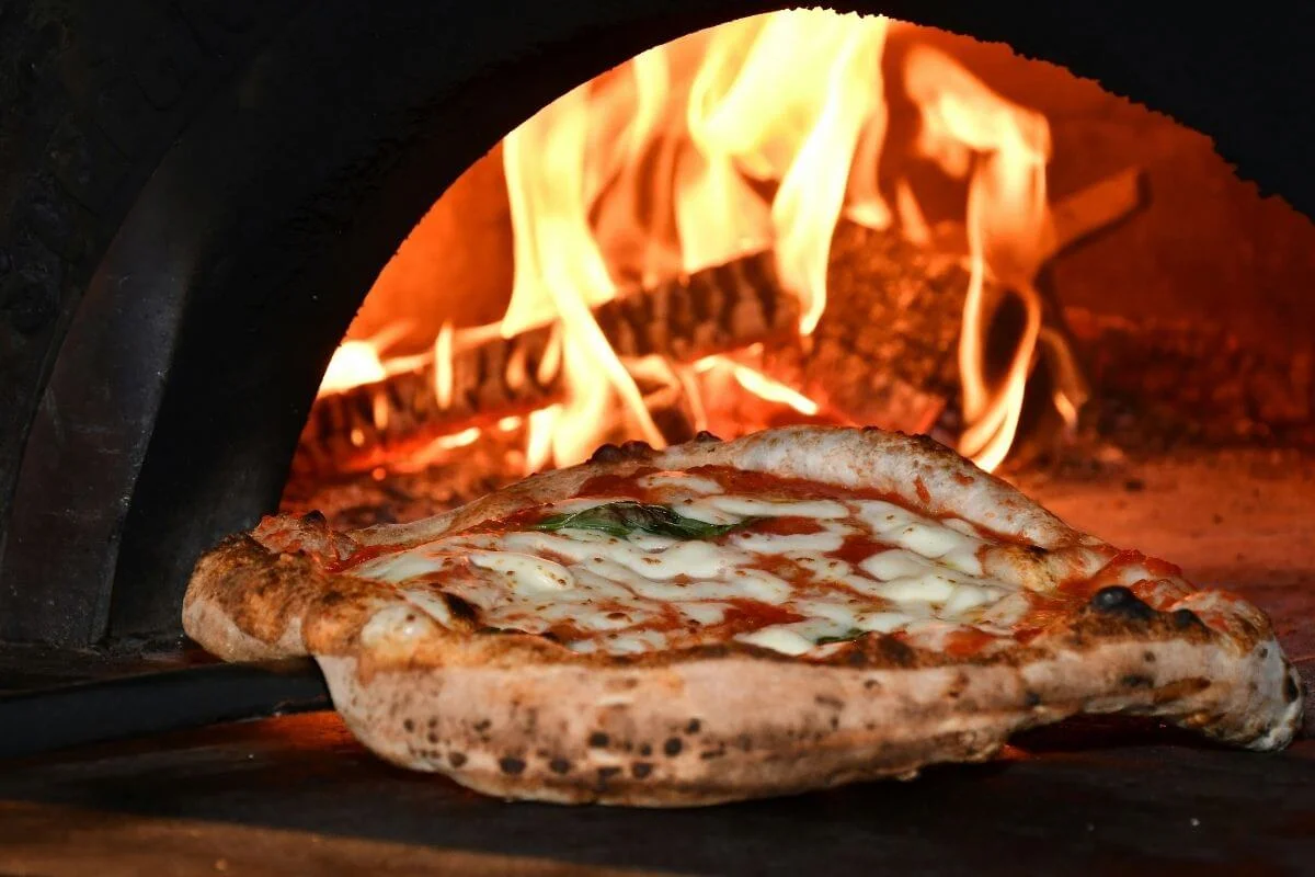 Close-up of a Neapolitan-style Margherita pizza with bubbly crust and melted cheese baking inside a wood-fired brick oven, with bright orange flames in the background.
