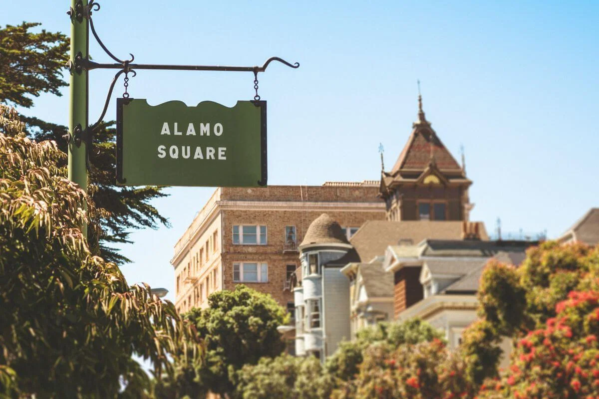 Green hanging sign reading “Alamo Square” in the foreground with leafy trees and classic San Francisco apartment and Victorian buildings softly blurred in the background under a clear blue sky.