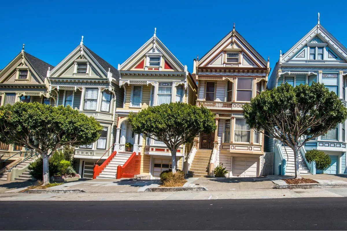 Row of brightly colored Victorian Painted Ladies houses behind neatly trimmed street trees on a steep San Francisco hill under a clear blue sky.