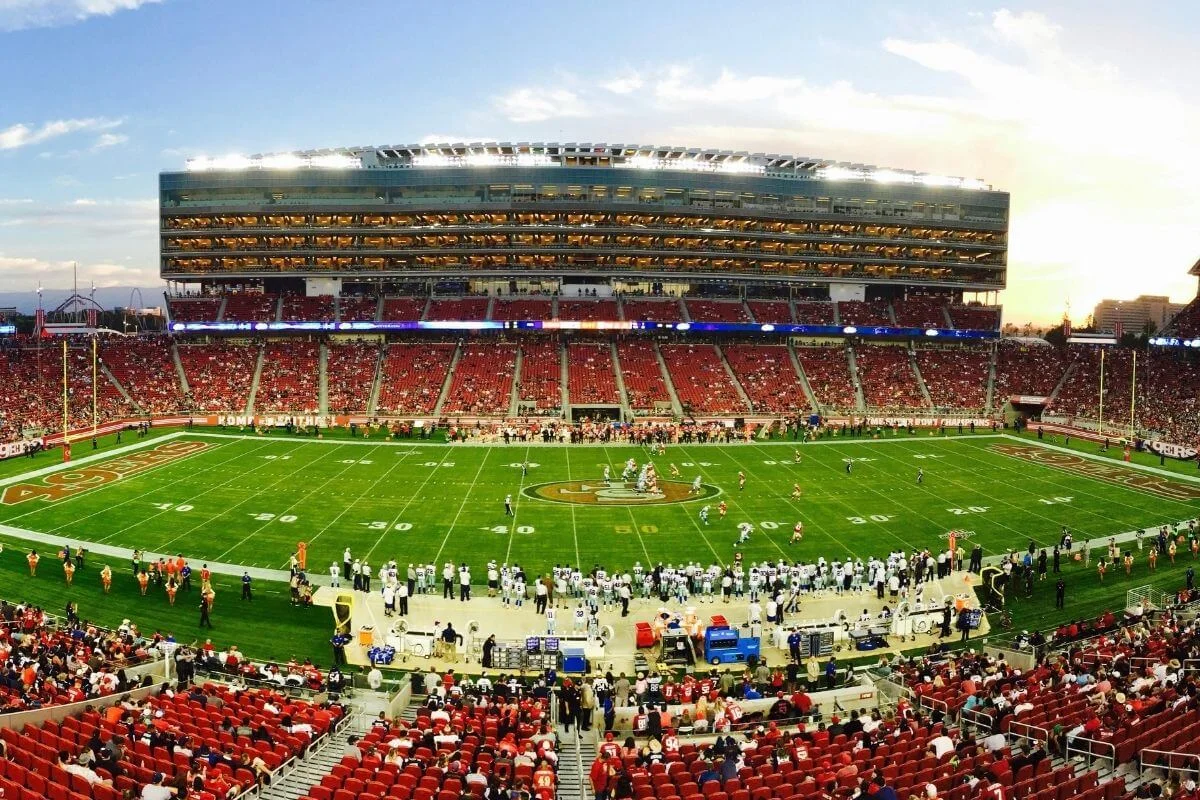 Wide view of a packed football stadium during a game, with players lined up on the field and fans filling red seats under stadium lights at sunset.
