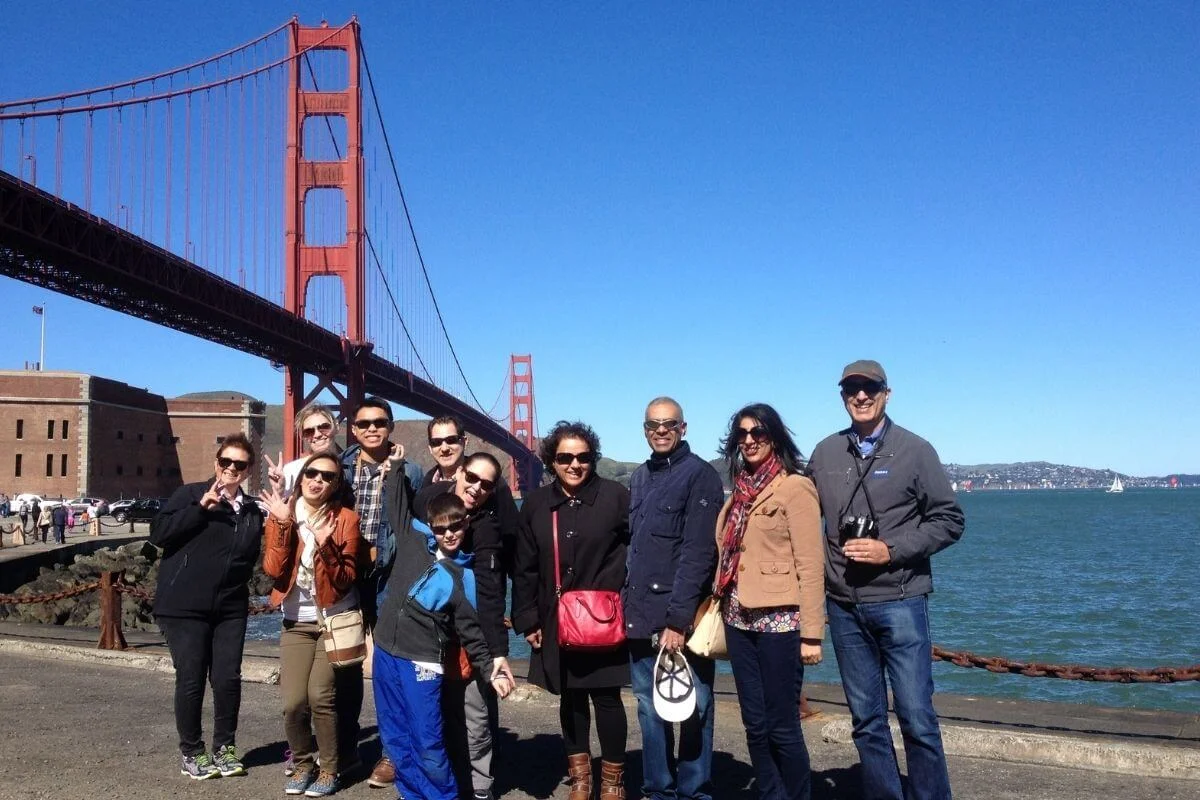 A group of people smiling and posing for a photo on a sunny day near the waterfront in San Francisco, with the Golden Gate Bridge and blue sky visible in the background.