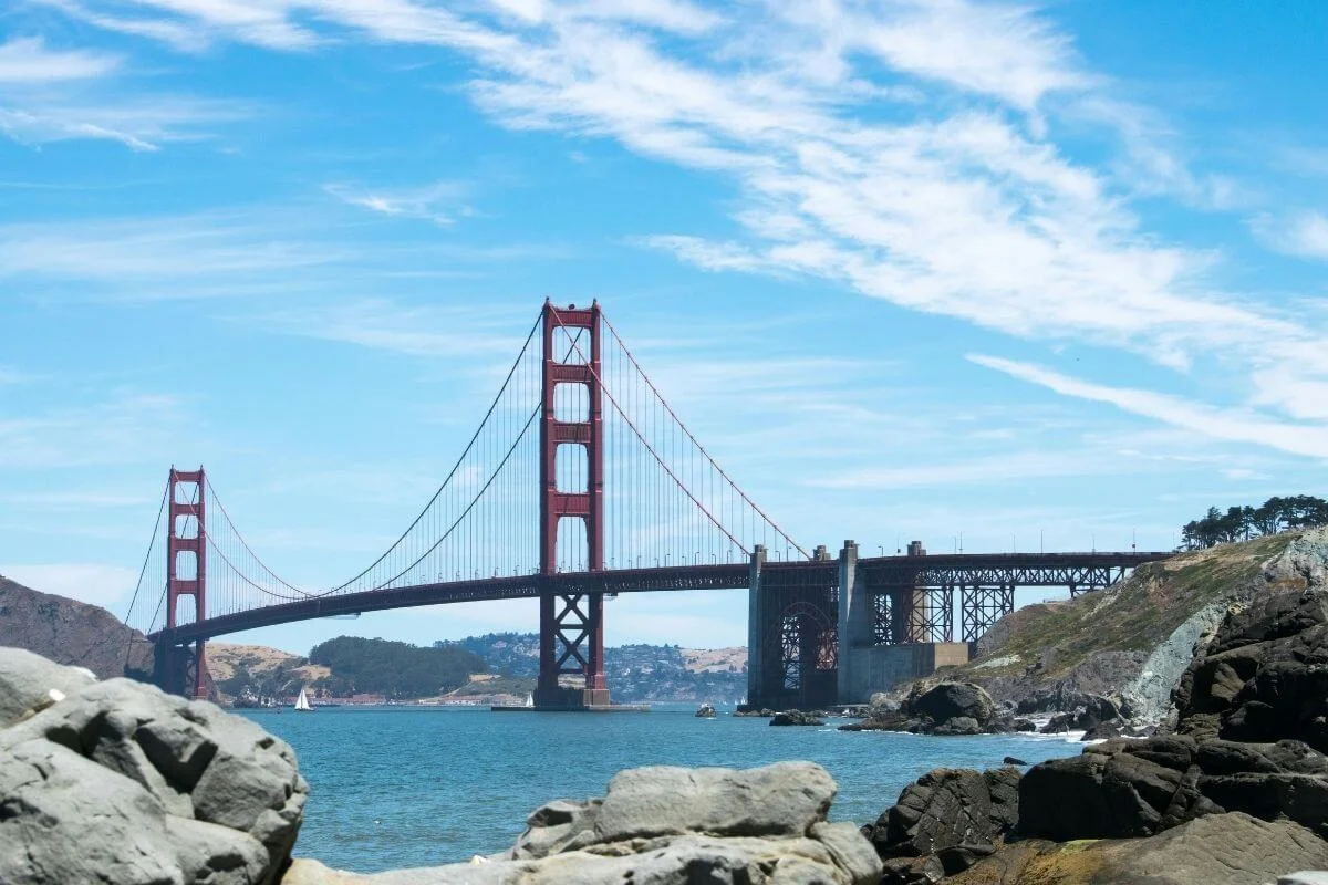 The Golden Gate Bridge spans the blue water of San Francisco Bay, viewed from a rocky coast with hills in the background under a bright sky.