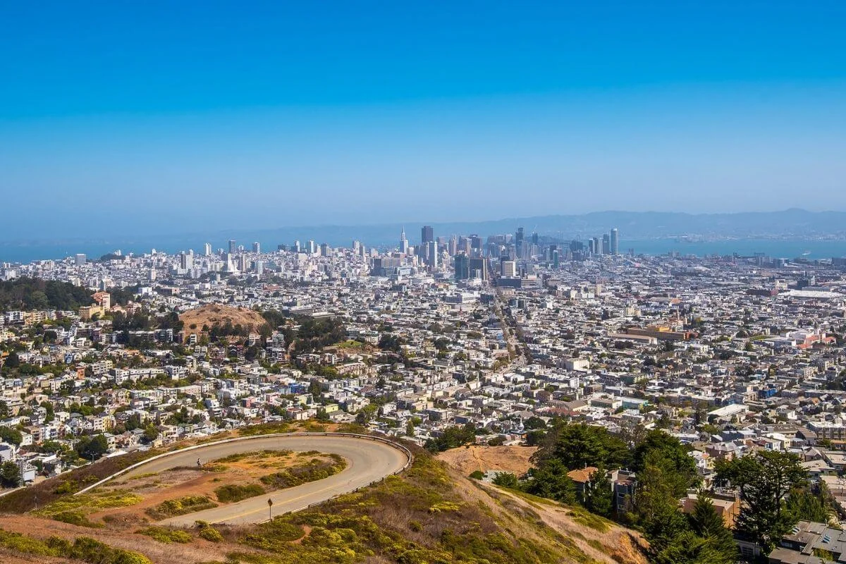 Panoramic view of San Francisco with the downtown skyline in the distance, seen from a hilltop overlook with a winding road in the foreground and the bay beyond.