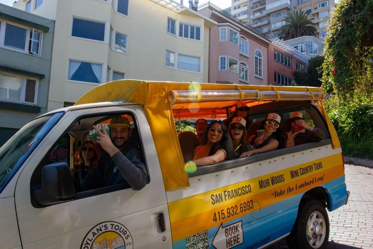 A cheerful group of people sit in a yellow and white open-air tour van labeled for San Francisco, smiling and posing for the camera as the driver flashes a peace sign on a sunny day in a residential city neighborhood.