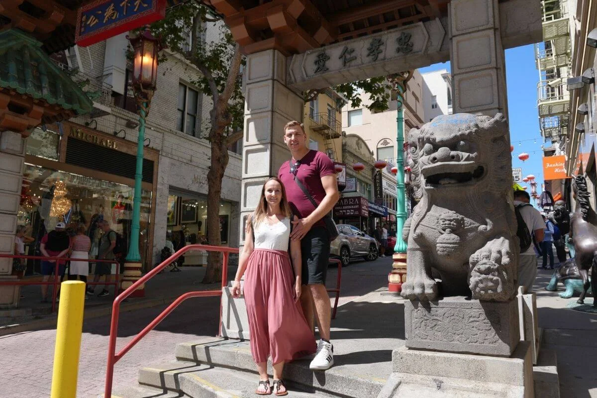A couple stands on the steps beside a stone lion statue under the ornate Chinatown gate in San Francisco, with shops, lanterns, and other pedestrians visible along the busy street behind them.