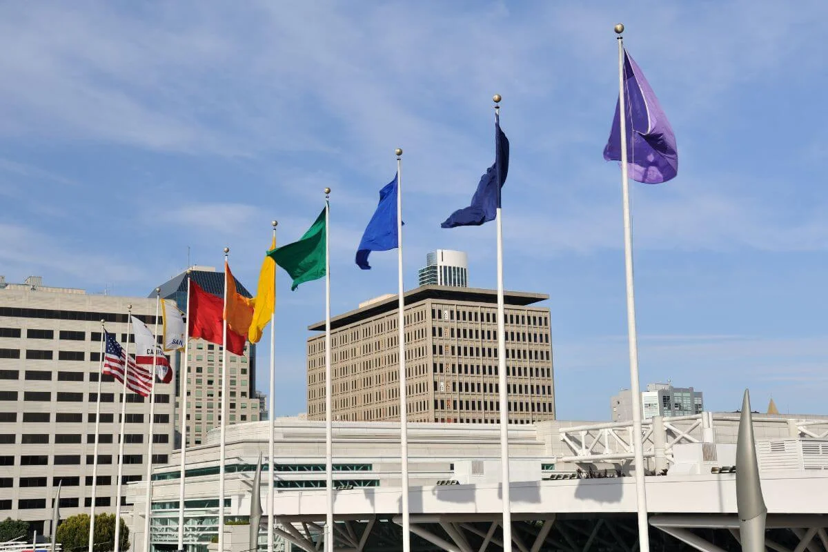 A row of colorful flags, including the U.S. and California flags, wave on tall poles in front of modern office buildings and a convention center under a bright blue sky.