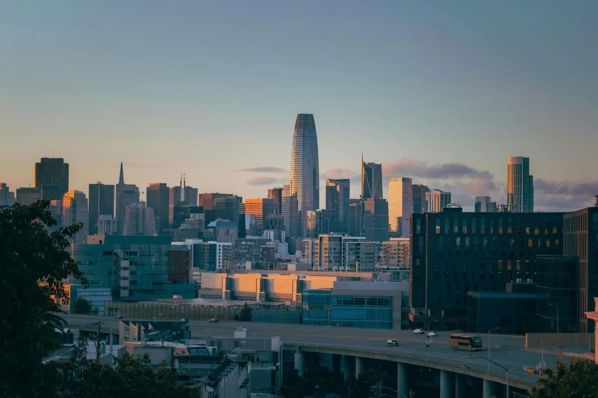View of the San Francisco skyline at sunset, with the tall Salesforce Tower and surrounding high-rise buildings glowing in warm light above a freeway and city rooftops.