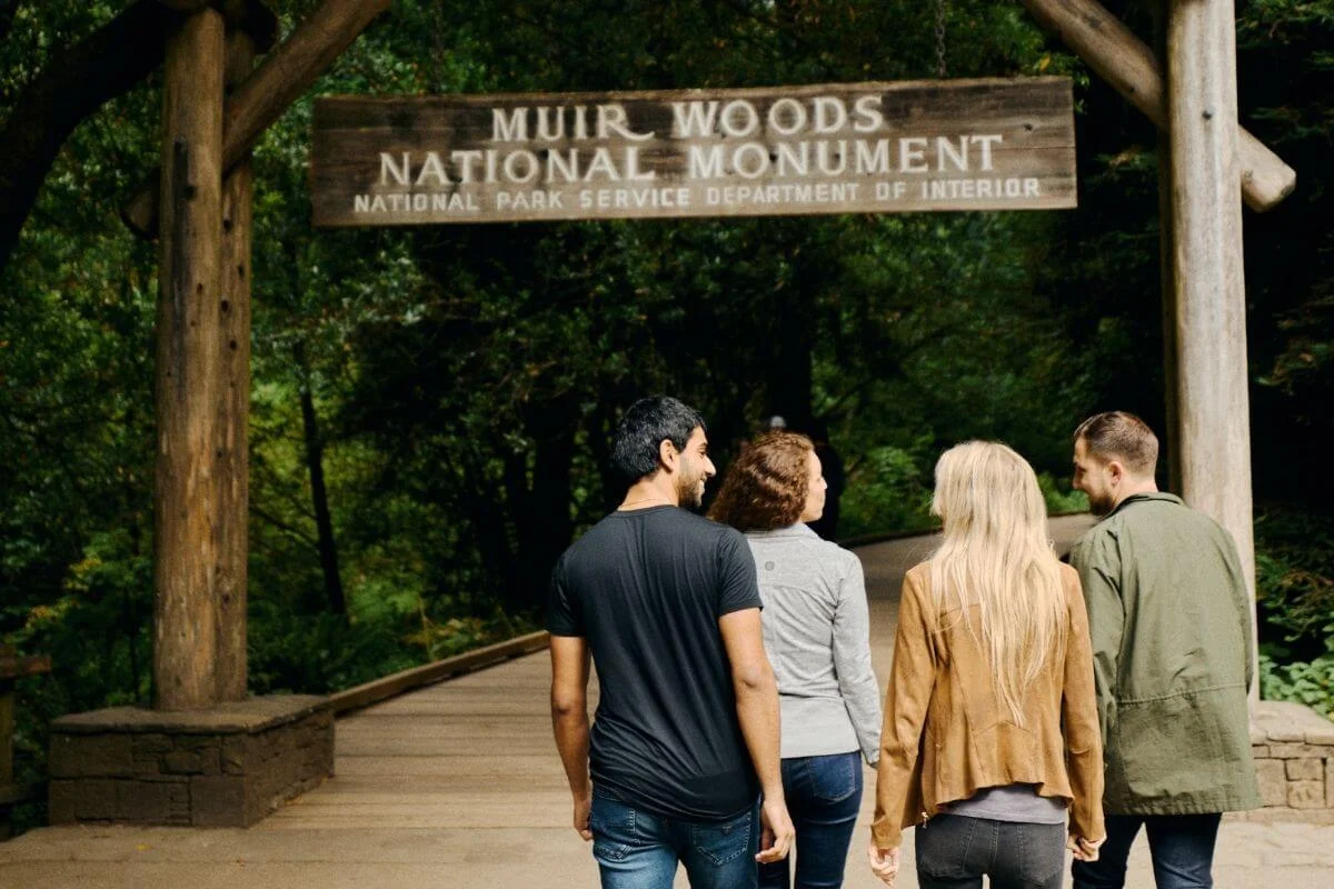 Four visitors walk under the Muir Woods National Monument entrance sign along a wooden path surrounded by redwood forest.
