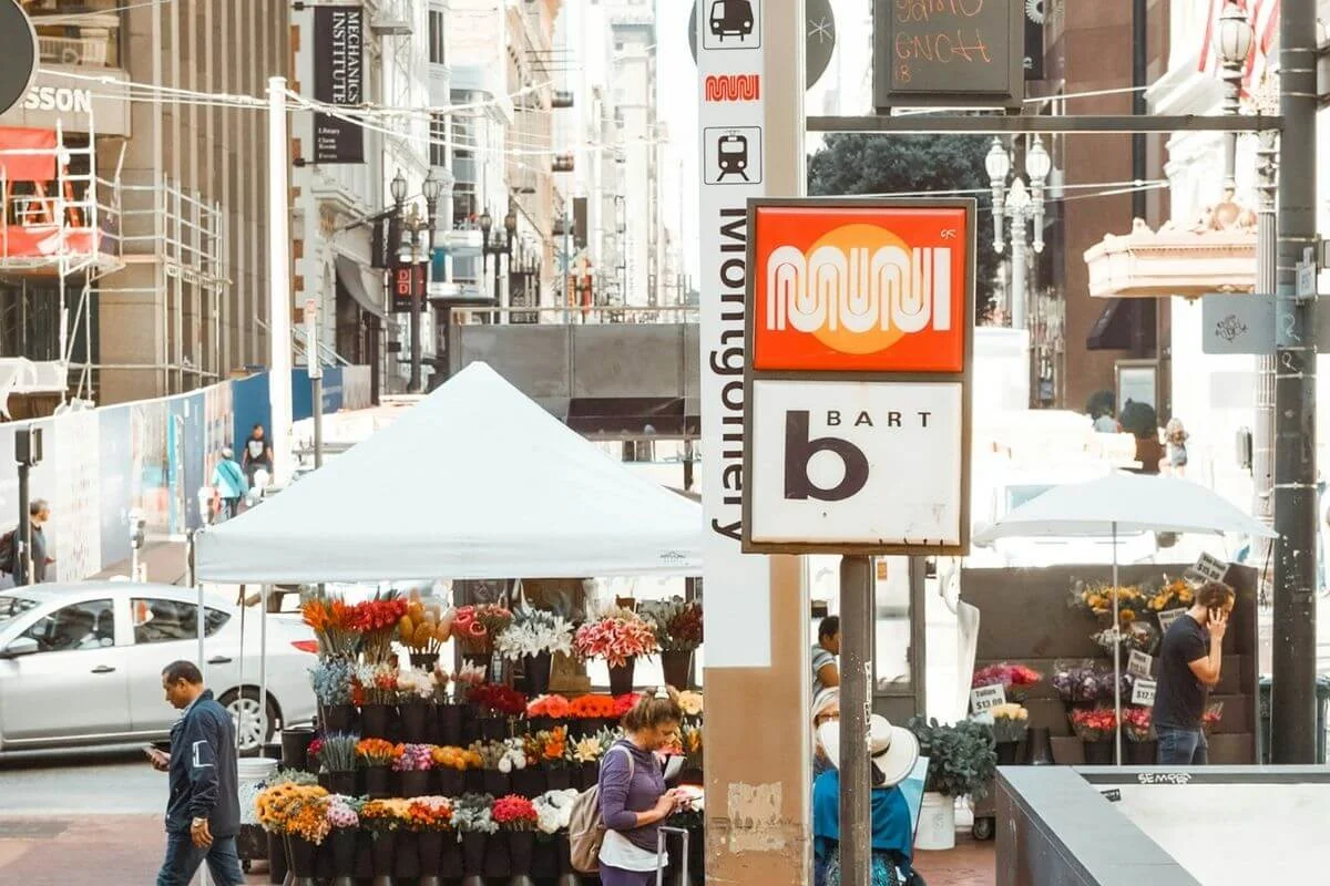 Busy downtown San Francisco street with a BART and Muni station sign in the foreground, flower stalls under white tents, pedestrians walking and shopping, and city buildings lining the background.