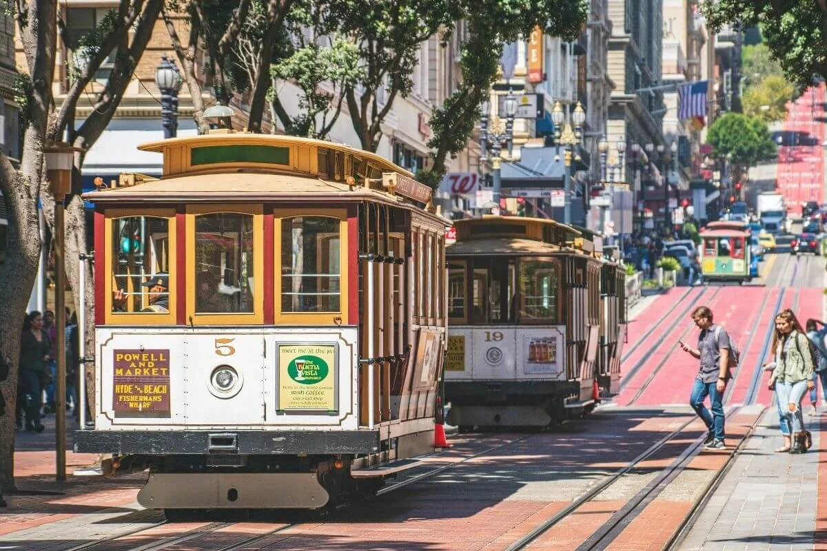 Historic San Francisco cable cars travel along tracks on a busy Powell Street, with pedestrians crossing in front, trees lining the sidewalk, and shops and traffic visible in the background.