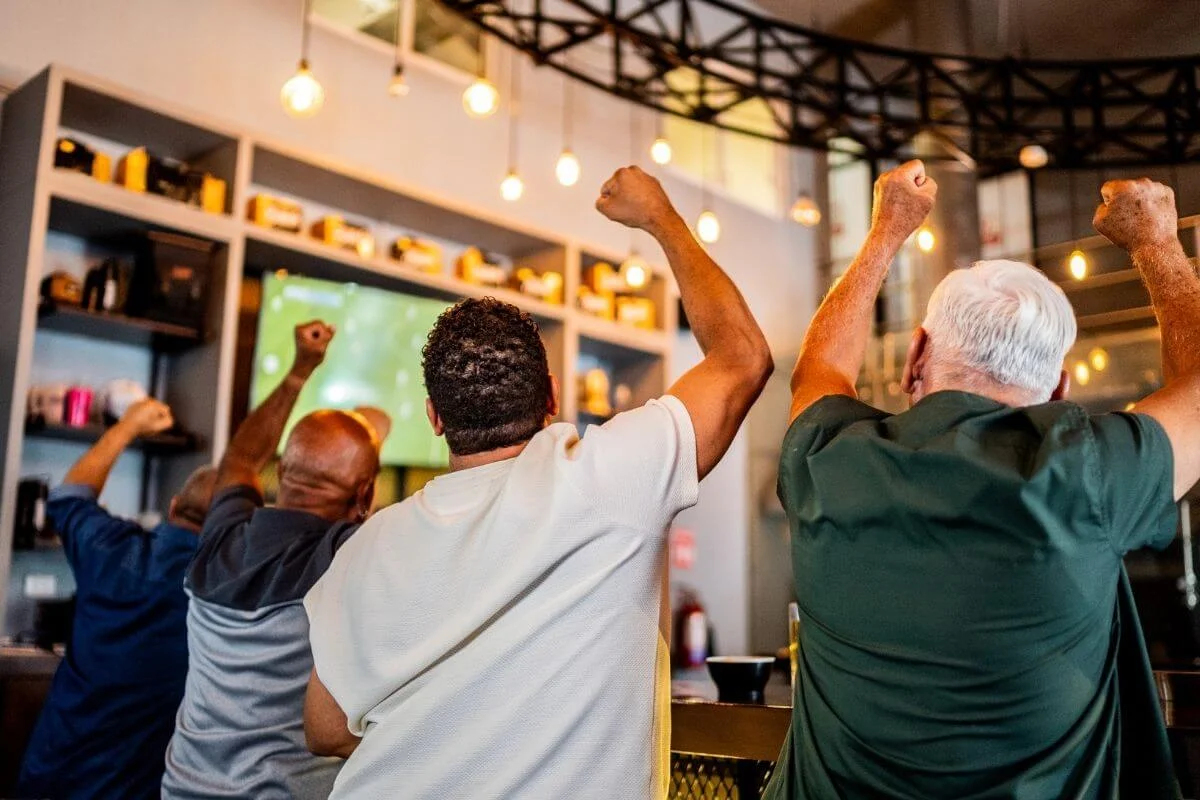 Group of people seen from behind raising their fists in celebration while watching a game on a TV inside a bar with hanging lights.