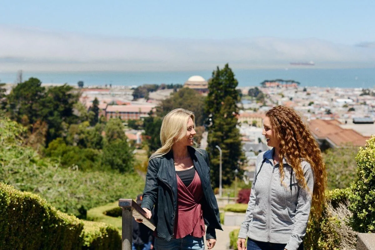 Two women walk and talk along a garden path on a hillside with manicured hedges, with a panoramic view of San Francisco, the Palace of Fine Arts dome, and the bay in the background.