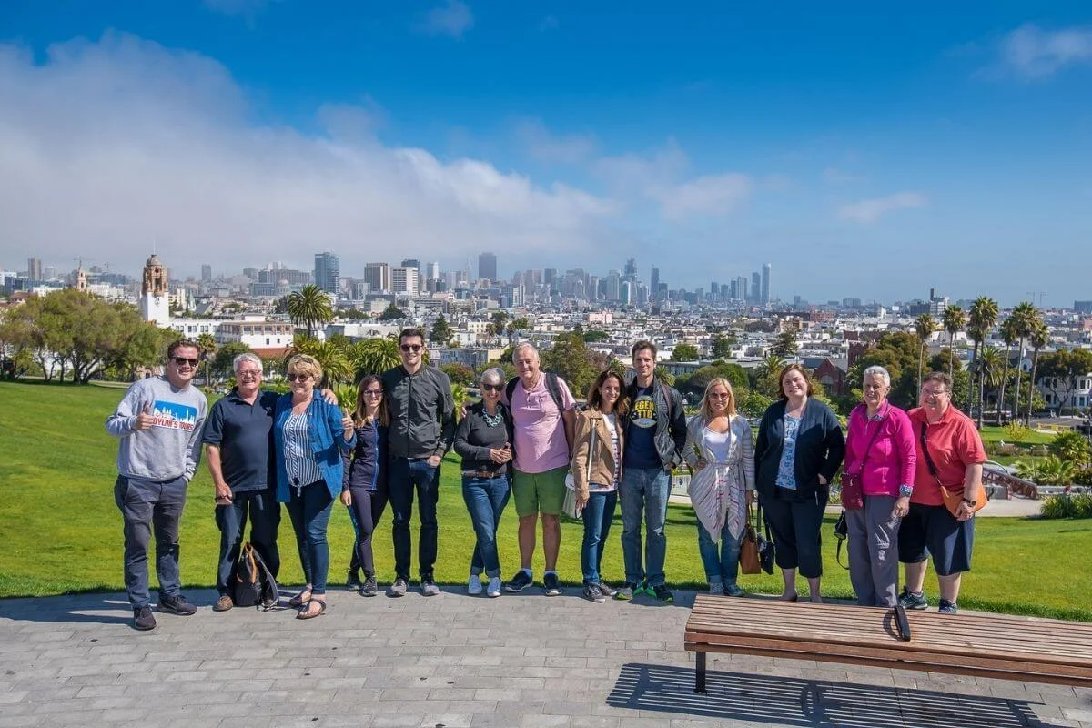 A group of people stand in a line posing for a photo on a paved path in Dolores Park, with green lawns, palm trees, and the San Francisco skyline and light fog in the background under a bright blue sky.