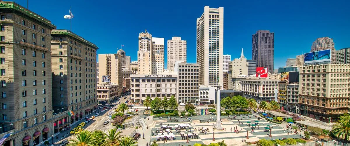 A wide daytime view of San Francisco’s Union Square, showing the central monument, trees, outdoor seating areas, and people walking through the plaza, surrounded by tall city buildings under a clear blue sky.