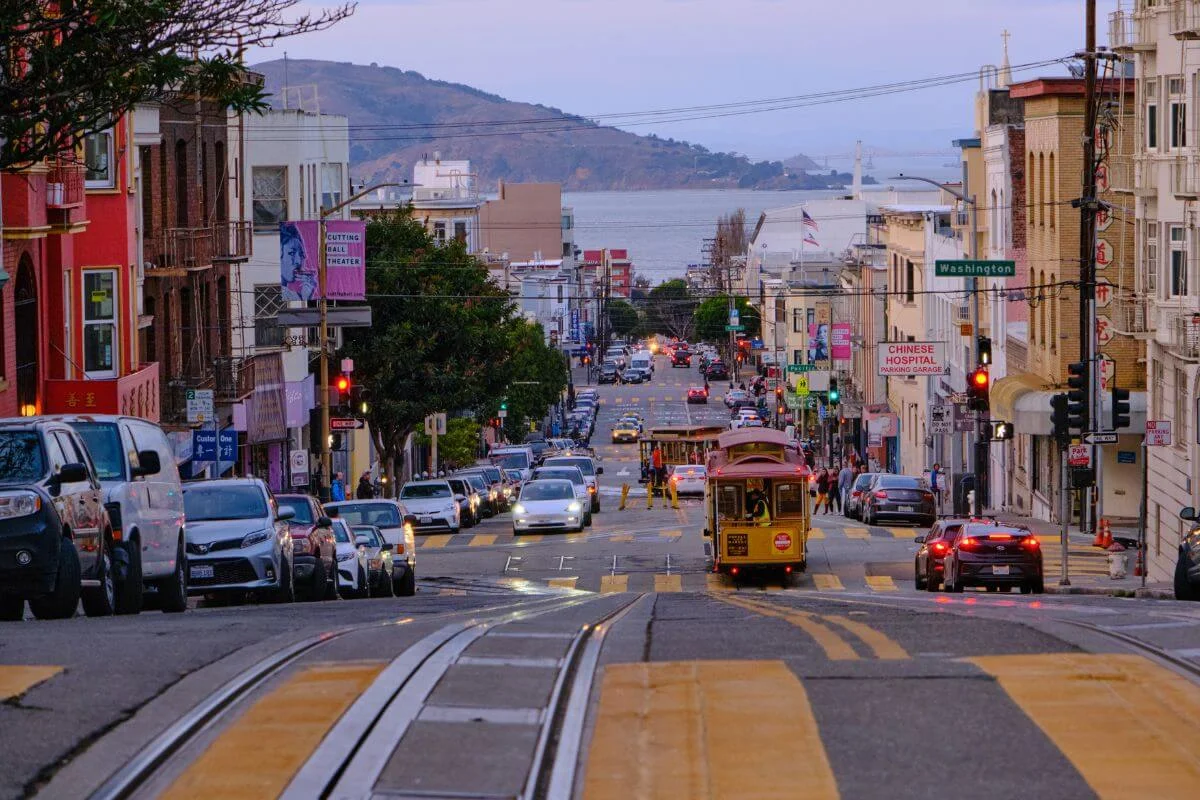 A classic San Francisco cable car traveling down a busy city street with cars, storefronts, and pedestrians on both sides, rail tracks in the foreground, and the bay and hills visible in the distance.
