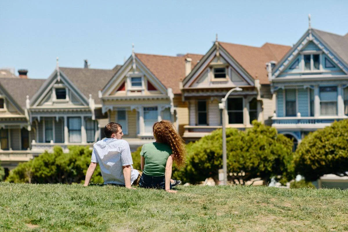 Two people sitting on a grassy hill at Alamo Square with the Painted Ladies Victorian houses visible in the background on a sunny day in San Francisco.